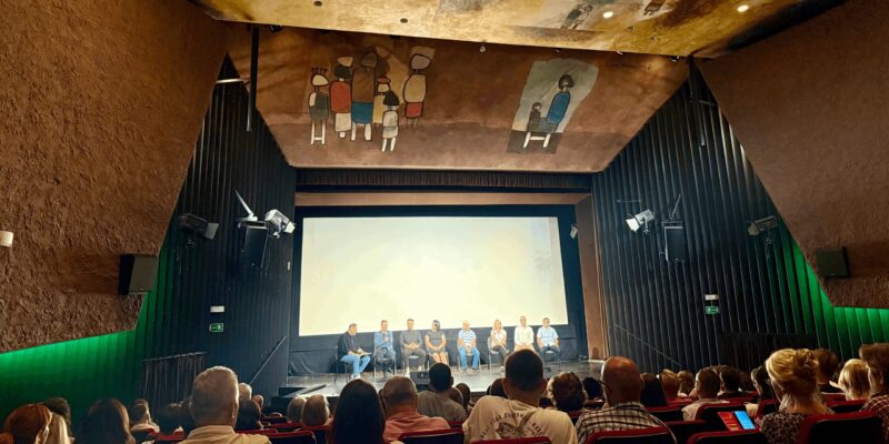 Publiczność wypełnia salę Oświęcimskiego Centrum Kultury podczas premiery filmu „Rok 2020” Piotra Struczyka; na scenie przed ekranem siedzą twórcy i aktorzy, a sufit zdobi charakterystyczne malowidło z postaciami w styluPubliczność wypełnia salę Oświęcimskiego Centrum Kultury podczas premiery filmu „Rok 2020” Piotra Struczyka; na scenie przed ekranem siedzą twórcy i aktorzy, a sufit zdobi charakterystyczne malowidło z postaciami w stylu nawiązującym do sztuki współczesnej. nawiązującym do sztuki współczesnej.