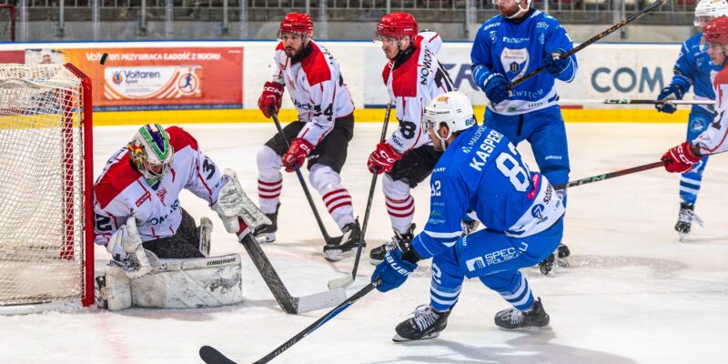 Zawodnik Unii Oświęcim w niebieskim stroju próbuje zdobyć gola w meczu z Cracovią. Bramkarz gospodarzy broni strzał, a kilku hokeistów w biało-czerwonych strojach blokuje dostęp do bramki podczas ligowego spotkania TAURON Hokej Ligi w Krakowie.