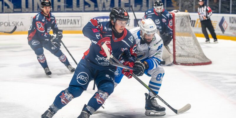 Zawodnik KH Energi Toruń walczy o krążek z hokeistą Unii Oświęcim podczas meczu w Hali Lodowej przy ulicy Chemików 4. W tle widoczna bramka, bramkarz i kibice z transparentem „KS Unia Oświęcim”.