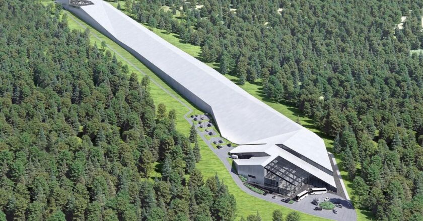 Wizualizacja nowoczesnej hali narciarskiej w Babicy w gminie Wadowice, otoczonej lasem. Długi, kryty stok o imponującej długości 650 metrów ciągnie się wśród drzew, prowadząc do przeszklonego budynku z parkingiem i autobusami przed wejściem.