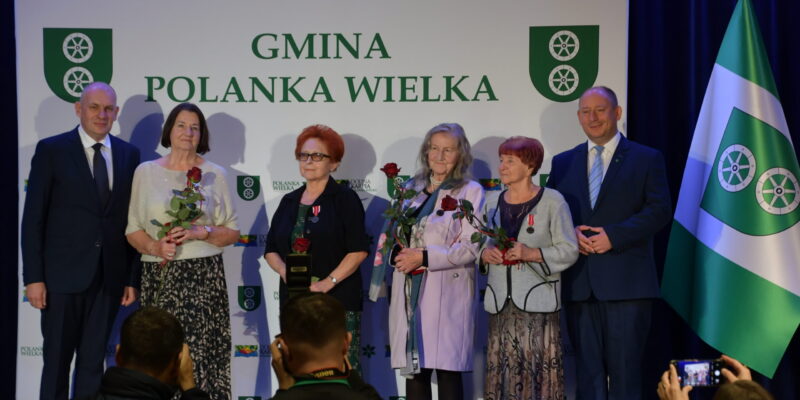 Uhonorowane mieszkanki Polanki Wielkiej podczas obchodów 700-lecia gminy. Kobiety trzymają róże i medale, stojąc obok władz samorządowych na tle ścianki z napisem „Gmina Polanka Wielka”.
