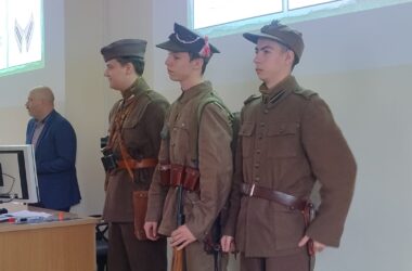 Trzech młodych mężczyzn ubranych w rekonstrukcje mundurów wojskowych II Rzeczypospolitej stoi w szeregu podczas wykładu w Małopolskiej Uczelni Państwowej. Obok nich, przy biurku z komputerem, stoi prowadzący prelekcję. W tle na ekranie wyświetlane są slajdy dotyczące umundurowania historycznego.