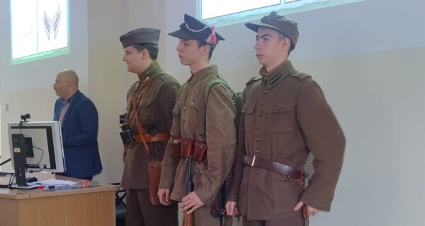 Trzech młodych mężczyzn ubranych w rekonstrukcje mundurów wojskowych II Rzeczypospolitej stoi w szeregu podczas wykładu w Małopolskiej Uczelni Państwowej. Obok nich, przy biurku z komputerem, stoi prowadzący prelekcję. W tle na ekranie wyświetlane są slajdy dotyczące umundurowania historycznego.