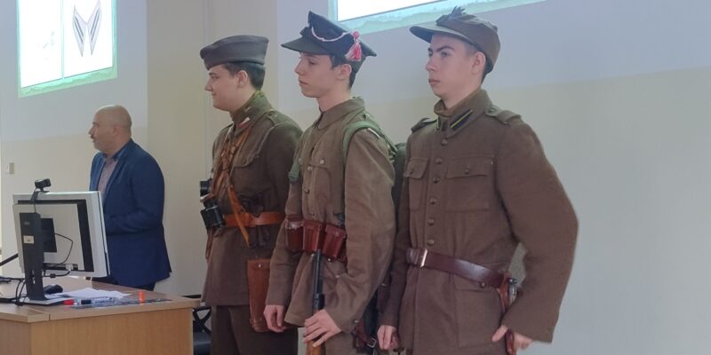 Trzech młodych mężczyzn ubranych w rekonstrukcje mundurów wojskowych II Rzeczypospolitej stoi w szeregu podczas wykładu w Małopolskiej Uczelni Państwowej. Obok nich, przy biurku z komputerem, stoi prowadzący prelekcję. W tle na ekranie wyświetlane są slajdy dotyczące umundurowania historycznego.