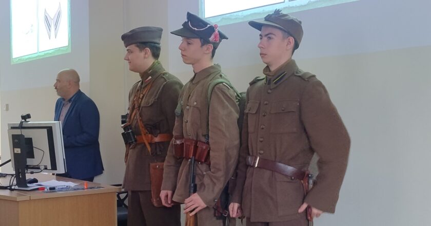 Trzech młodych mężczyzn ubranych w rekonstrukcje mundurów wojskowych II Rzeczypospolitej stoi w szeregu podczas wykładu w Małopolskiej Uczelni Państwowej. Obok nich, przy biurku z komputerem, stoi prowadzący prelekcję. W tle na ekranie wyświetlane są slajdy dotyczące umundurowania historycznego.