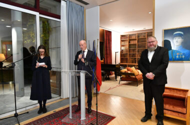 Ambasador Francji Étienne de Poncins stojący przy mównicy podczas uroczystości w Rezydencji Ambasadora Francji w Warszawie. Po lewej stronie stoi tłumaczka, a po prawej dyrektor Muzeum Auschwitz dr Piotr M. A. Cywiński. W tle widoczne są regały z książkami, fortepian oraz wiszący na ścianie portret przedstawiciela francuskiej armii.