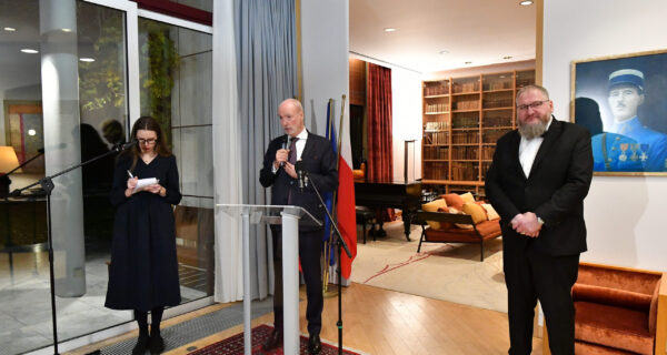 Ambasador Francji Étienne de Poncins stojący przy mównicy podczas uroczystości w Rezydencji Ambasadora Francji w Warszawie. Po lewej stronie stoi tłumaczka, a po prawej dyrektor Muzeum Auschwitz dr Piotr M. A. Cywiński. W tle widoczne są regały z książkami, fortepian oraz wiszący na ścianie portret przedstawiciela francuskiej armii.