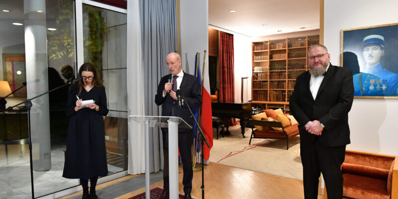 Ambasador Francji Étienne de Poncins stojący przy mównicy podczas uroczystości w Rezydencji Ambasadora Francji w Warszawie. Po lewej stronie stoi tłumaczka, a po prawej dyrektor Muzeum Auschwitz dr Piotr M. A. Cywiński. W tle widoczne są regały z książkami, fortepian oraz wiszący na ścianie portret przedstawiciela francuskiej armii.