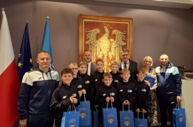 Grupa młodych piłkarzy ze Szkoły Podstawowej nr 5 w Oświęcimiu pozuje do pamiątkowego zdjęcia z prezydentem Oświęcimia Januszem Chwierutem, jego zastępcą Krzysztofem Kanią, dyrektorką szkoły Renatą Biesik-Myśliwiec oraz trenerami. Chłopcy ubrani są w jednakowe sportowe bluzy i trzymają w rękach niebieskie torby z herbem miasta. Za grupą widać dużą stylizowaną grafikę orła oraz flagi Polski, UE i Oświęcimia. Zdjęcie wykonane w reprezentacyjnej sali urzędu.
