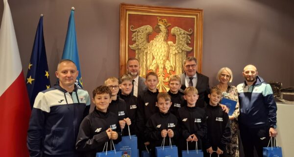 Grupa młodych piłkarzy ze Szkoły Podstawowej nr 5 w Oświęcimiu pozuje do pamiątkowego zdjęcia z prezydentem Oświęcimia Januszem Chwierutem, jego zastępcą Krzysztofem Kanią, dyrektorką szkoły Renatą Biesik-Myśliwiec oraz trenerami. Chłopcy ubrani są w jednakowe sportowe bluzy i trzymają w rękach niebieskie torby z herbem miasta. Za grupą widać dużą stylizowaną grafikę orła oraz flagi Polski, UE i Oświęcimia. Zdjęcie wykonane w reprezentacyjnej sali urzędu.