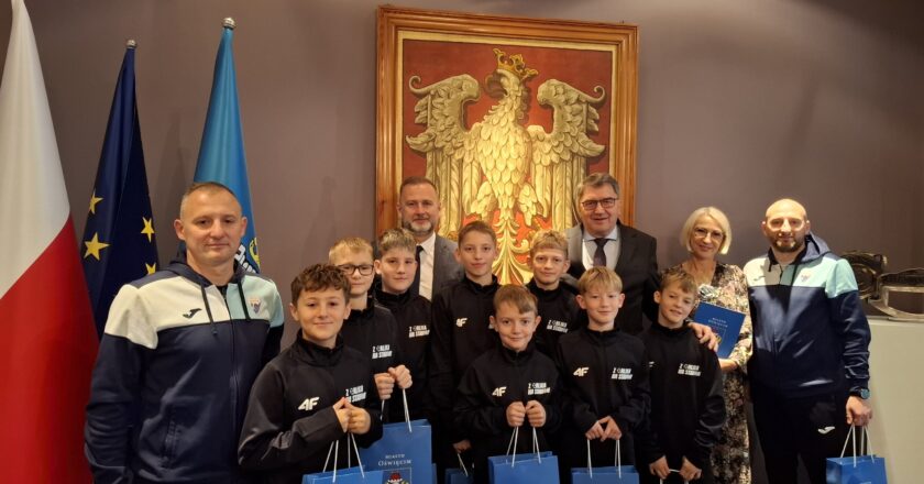 Grupa młodych piłkarzy ze Szkoły Podstawowej nr 5 w Oświęcimiu pozuje do pamiątkowego zdjęcia z prezydentem Oświęcimia Januszem Chwierutem, jego zastępcą Krzysztofem Kanią, dyrektorką szkoły Renatą Biesik-Myśliwiec oraz trenerami. Chłopcy ubrani są w jednakowe sportowe bluzy i trzymają w rękach niebieskie torby z herbem miasta. Za grupą widać dużą stylizowaną grafikę orła oraz flagi Polski, UE i Oświęcimia. Zdjęcie wykonane w reprezentacyjnej sali urzędu.