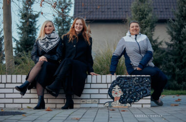 Trzy kobiety biorące udział w projekcie „Mozaiki – Miasto Pracowni” siedzą na białej ławce obok jednej z nowych mozaik przedstawiających kobietę z rozwianymi włosami; po prawej stronie widoczna jest mozaika, po lewej autorki uśmiechające się do obiektywu