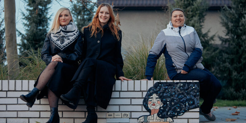 Trzy kobiety biorące udział w projekcie „Mozaiki – Miasto Pracowni” siedzą na białej ławce obok jednej z nowych mozaik przedstawiających kobietę z rozwianymi włosami; po prawej stronie widoczna jest mozaika, po lewej autorki uśmiechające się do obiektywu