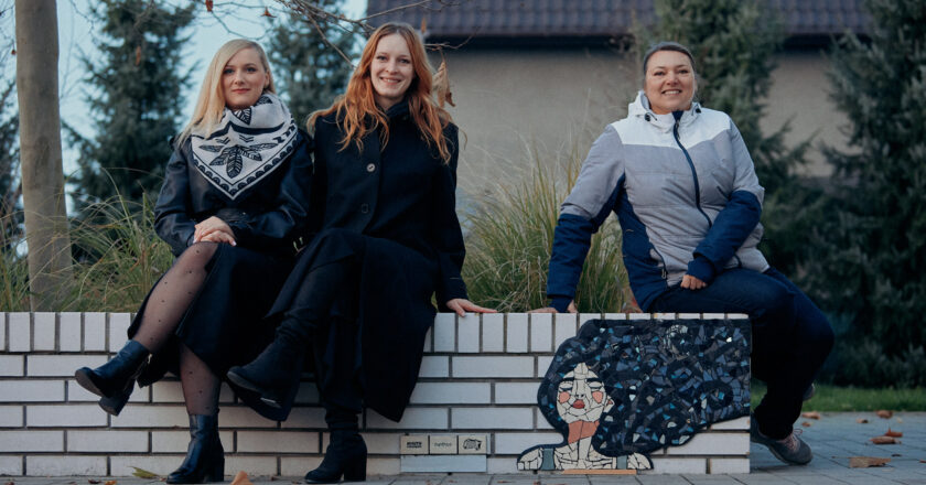 Trzy kobiety biorące udział w projekcie „Mozaiki – Miasto Pracowni” siedzą na białej ławce obok jednej z nowych mozaik przedstawiających kobietę z rozwianymi włosami; po prawej stronie widoczna jest mozaika, po lewej autorki uśmiechające się do obiektywu