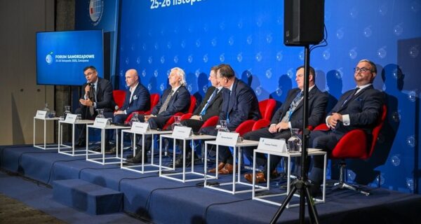 Paneliści siedzący na scenie podczas Forum Samorządowego w Zakopanem; w tle niebieska ściana z logo wydarzenia i datą „25–26 listopada 2025”, Radosław Włoszek po lewej zabiera głos do mikrofonu.