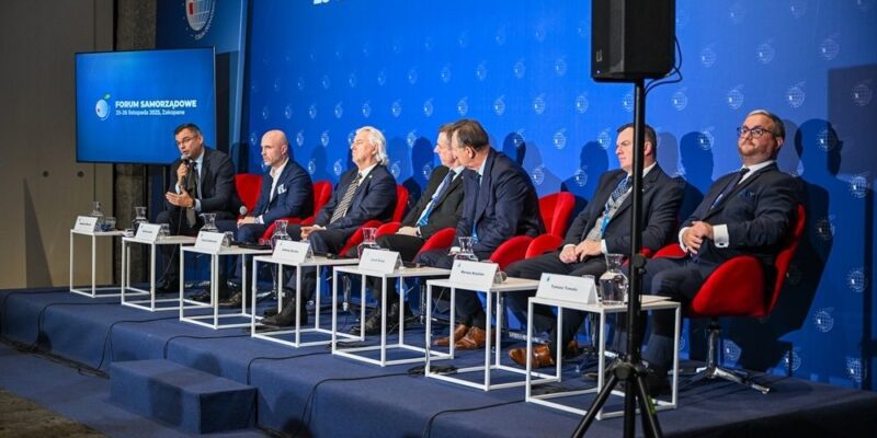Paneliści siedzący na scenie podczas Forum Samorządowego w Zakopanem; w tle niebieska ściana z logo wydarzenia i datą „25–26 listopada 2025”, Radosław Włoszek po lewej zabiera głos do mikrofonu.