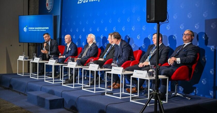 Paneliści siedzący na scenie podczas Forum Samorządowego w Zakopanem; w tle niebieska ściana z logo wydarzenia i datą „25–26 listopada 2025”, Radosław Włoszek po lewej zabiera głos do mikrofonu.