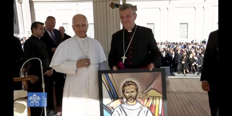 Biskup Roman Pindel przekazuje papieżowi Leonowi XIV obraz z wizerunkiem bł. Michała Tomaszka podczas audiencji na Placu św. Piotra; obaj stoją obok siebie na podium, w tle widać uczestników audiencji.