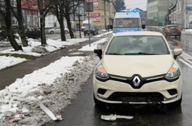 Białe Renault z uszkodzoną przednią częścią stoi na mokrej jezdni w Kętach po potrąceniu pieszej; za samochodem widać radiowóz policji z włączonymi sygnałami, a wokół leży śnieg i fragmenty uszkodzonych elementów pojazdu.