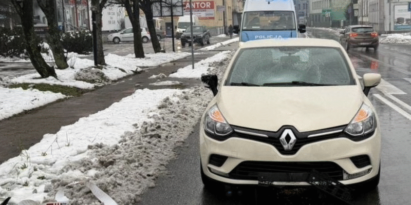 Białe Renault z uszkodzoną przednią częścią stoi na mokrej jezdni w Kętach po potrąceniu pieszej; za samochodem widać radiowóz policji z włączonymi sygnałami, a wokół leży śnieg i fragmenty uszkodzonych elementów pojazdu.