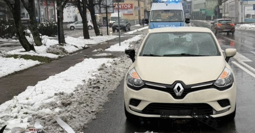 Białe Renault z uszkodzoną przednią częścią stoi na mokrej jezdni w Kętach po potrąceniu pieszej; za samochodem widać radiowóz policji z włączonymi sygnałami, a wokół leży śnieg i fragmenty uszkodzonych elementów pojazdu.