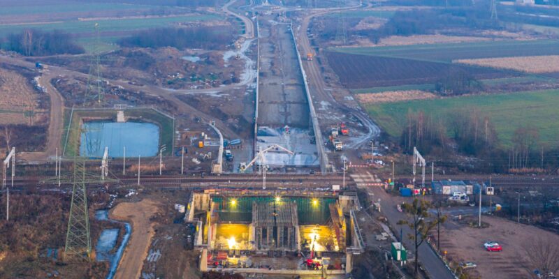 Widok z drona na budowę obwodnicy Oświęcimia: powstający tunel pod torami kolejowymi oraz długi odcinek nowej drogi przebiegającej przez tereny rolnicze w rejonie Brzezinki.