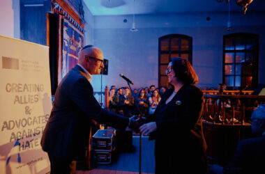 Uroczystość jubileuszowa muzeum żydowskiego w Oświęcimiu – dwie osoby podają sobie rękę na scenie podczas wręczania nagrody „Fighting Hatred”, w tle publiczność i podświetlone wnętrze zabytkowej synagogi.