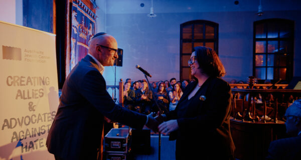 Uroczystość jubileuszowa muzeum żydowskiego w Oświęcimiu – dwie osoby podają sobie rękę na scenie podczas wręczania nagrody „Fighting Hatred”, w tle publiczność i podświetlone wnętrze zabytkowej synagogi.