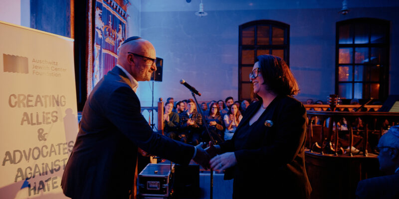 Uroczystość jubileuszowa muzeum żydowskiego w Oświęcimiu – dwie osoby podają sobie rękę na scenie podczas wręczania nagrody „Fighting Hatred”, w tle publiczność i podświetlone wnętrze zabytkowej synagogi.