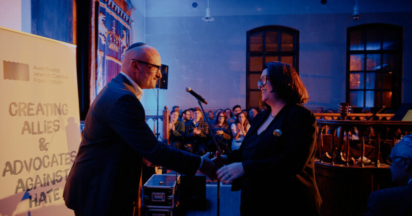 Uroczystość jubileuszowa muzeum żydowskiego w Oświęcimiu – dwie osoby podają sobie rękę na scenie podczas wręczania nagrody „Fighting Hatred”, w tle publiczność i podświetlone wnętrze zabytkowej synagogi.