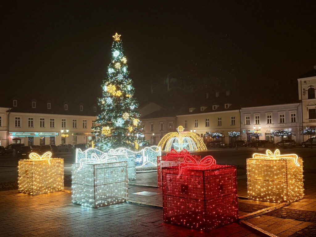 Świąteczna iluminacja na Rynku Głównym w Oświęcimiu: wysoka choinka, świetlne prezenty oraz dekoracyjna fontanna otoczone miejskimi kamienicami w wieczornej scenerii.
