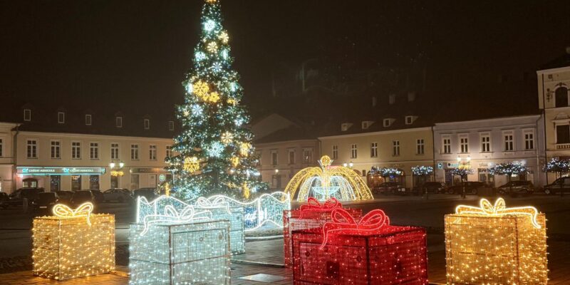 Świąteczna iluminacja na Rynku Głównym w Oświęcimiu: wysoka choinka, świetlne prezenty oraz dekoracyjna fontanna otoczone miejskimi kamienicami w wieczornej scenerii.