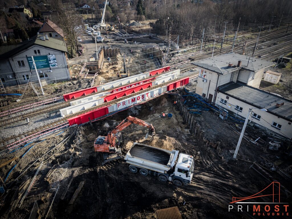 Budowa tunelu pod torami kolejowymi na ul. Wawelskiej w Bieruniu – widok z góry na konstrukcje odciążające firmy Primost Południe, prowadzone roboty ziemne, koparkę i ciężarówkę pracujące bezpośrednio pod czynnymi torami kolejowymi.