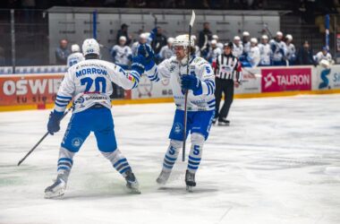 Dwaj hokeiści Unii Oświęcim w białoniebieskich strojach cieszą się po zdobyciu bramki podczas meczu, przy bandach widoczna ławka drużyny i sędzia na lodzie.