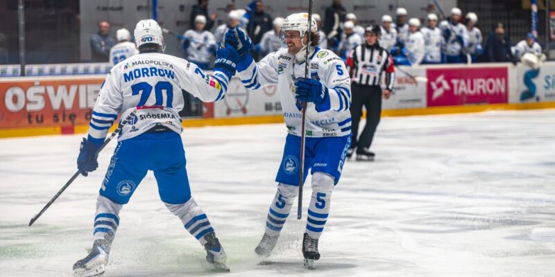 Dwaj hokeiści Unii Oświęcim w białoniebieskich strojach cieszą się po zdobyciu bramki podczas meczu, przy bandach widoczna ławka drużyny i sędzia na lodzie.