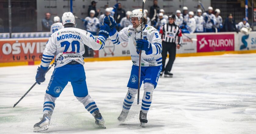 Dwaj hokeiści Unii Oświęcim w białoniebieskich strojach cieszą się po zdobyciu bramki podczas meczu, przy bandach widoczna ławka drużyny i sędzia na lodzie.