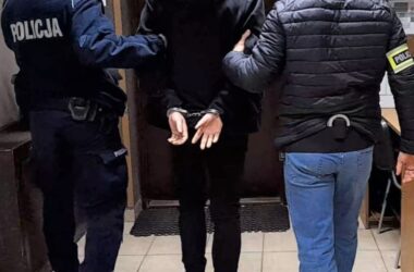 Zatrzymany mężczyzna w kajdankach prowadzony korytarzem przez dwóch policjantów – jednego w mundurze i jednego w ubraniu cywilnym z opaską „Policja”.