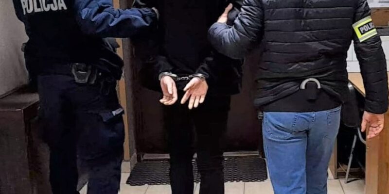 Zatrzymany mężczyzna w kajdankach prowadzony korytarzem przez dwóch policjantów – jednego w mundurze i jednego w ubraniu cywilnym z opaską „Policja”.