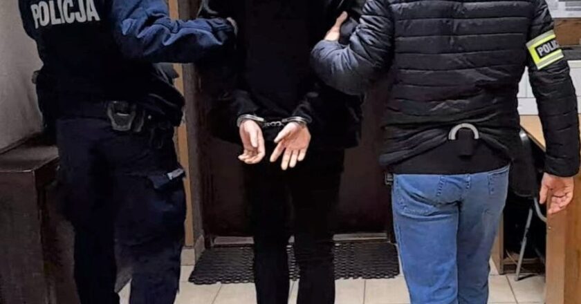 Zatrzymany mężczyzna w kajdankach prowadzony korytarzem przez dwóch policjantów – jednego w mundurze i jednego w ubraniu cywilnym z opaską „Policja”.