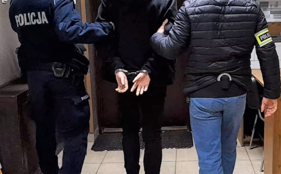 Zatrzymany mężczyzna w kajdankach prowadzony korytarzem przez dwóch policjantów – jednego w mundurze i jednego w ubraniu cywilnym z opaską „Policja”.
