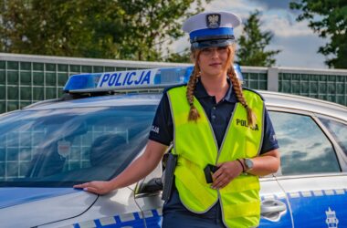 Policjantka ruchu drogowego w kamizelce odblaskowej stoi obok oznakowanego radiowozu policyjnego, w tle ogrodzenie i zieleń przy drodze.