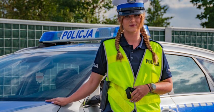 Policjantka ruchu drogowego w kamizelce odblaskowej stoi obok oznakowanego radiowozu policyjnego, w tle ogrodzenie i zieleń przy drodze.