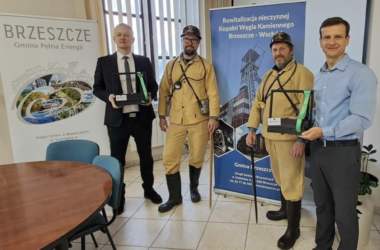 Delegacja w historycznych górniczych strojach wraz z przedstawicielami Urzędu Gminy Brzeszcze podczas barbórkowej wizyty, trzymająca pamiątkowe upominki na tle roll-upów o gminie i kopalni Jawiszowice