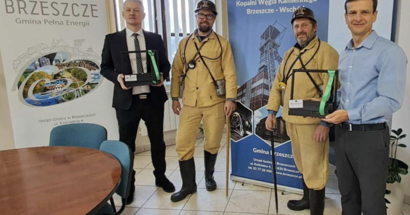 Delegacja w historycznych górniczych strojach wraz z przedstawicielami Urzędu Gminy Brzeszcze podczas barbórkowej wizyty, trzymająca pamiątkowe upominki na tle roll-upów o gminie i kopalni Jawiszowice