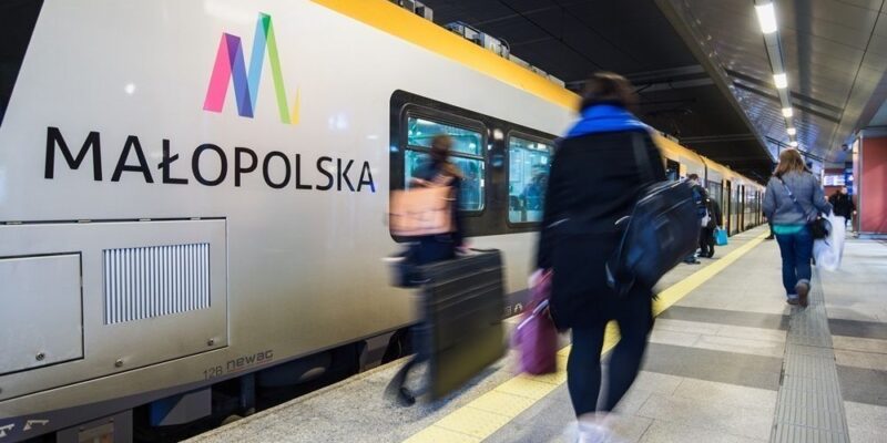 Pasażerowie wsiadający do pociągu Kolei Małopolskich na peronie podziemnego dworca, nowoczesny skład kolejowy z logo Małopolski, ruch i pośpiech w godzinach szczytu.