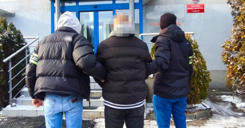 Funkcjonariusze Straży Granicznej prowadzą zatrzymanego obywatela Ukrainy do budynku Placówki Straży Granicznej w Krakowie; mężczyzna eskortowany przez dwóch mundurowych, widok od tyłu.