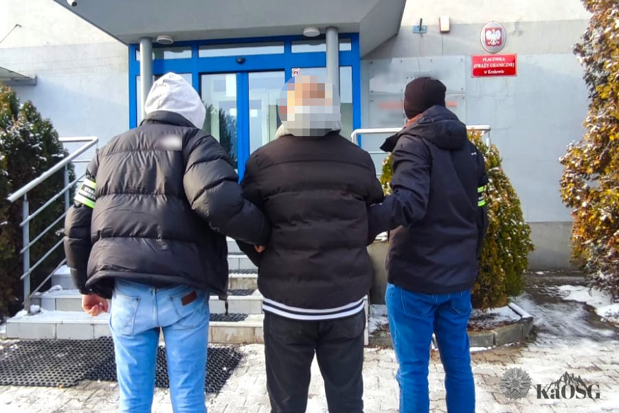 Funkcjonariusze Straży Granicznej prowadzą zatrzymanego obywatela Ukrainy do budynku Placówki Straży Granicznej w Krakowie; mężczyzna eskortowany przez dwóch mundurowych, widok od tyłu.