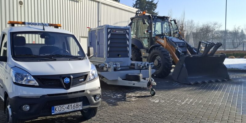 Nowy sprzęt zakupiony przez Miasto Oświęcim w ramach Programu Ochrony Ludności i Obrony Cywilnej – samochód do dystrybucji wody pitnej z przyczepą i motopompą oraz koparko-ładowarka MZUK, zaprezentowane na placu miejskim w słoneczny dzień.