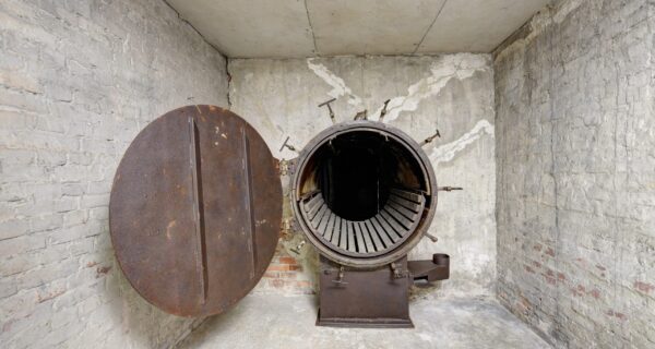 Wnętrze dawnej łaźni w Auschwitz II-Birkenau – otwarta metalowa komora dezynfekcyjna z okrągłymi drzwiami, widoczne ślady korozji i oryginalne elementy techniczne, surowe ceglane ściany i betonowa posadzka.