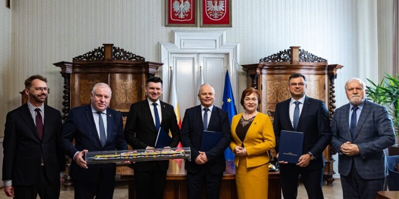 Podpisanie umowy na zakup nowych pociągów dla Małopolski – przedstawiciele Zarządu Województwa Małopolskiego, Kolei Małopolskich i spółki NEWAG S.A. pozują do zdjęcia w sali urzędowej, trzymając dokumenty kontraktowe i model pociągu.
